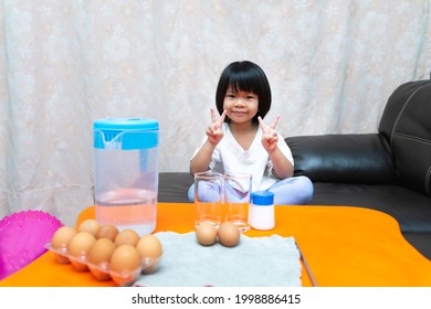Student sat on sofas waiting to learn experiment. Girl held up two fingers to take picture with sweet smile. Kid will conduct  sunken egg science experiment - the egg floats with water and salt water.