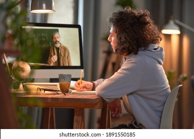 Student sitting at the table in front of computer monitor and listening to his teacher during online studying