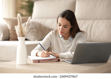 Student studying taking notes on notebook beside a laptop at home