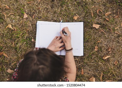 Student taking notes with a pen in a small notebook while sitting on the grass outdoors - Millennial girl handwriting lessons and information in the park