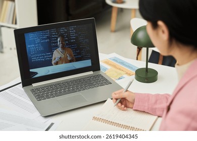 Student taking notes while watching an online language lesson on laptop in classroom with textbooks and study materials visible around. Engaging with diverse educational content
