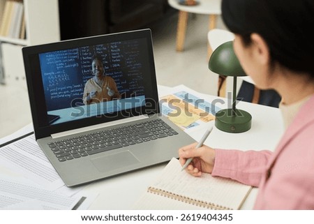 Student taking notes while watching an online language lesson on laptop in classroom with textbooks and study materials visible around. Engaging with diverse educational content