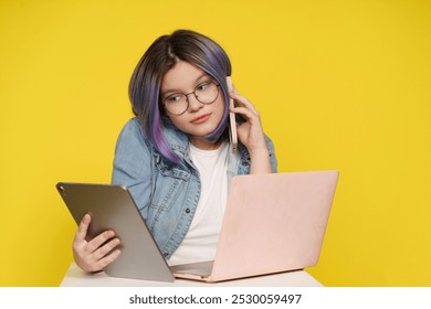 A student is talking on her cell phone while holding a tablet and a laptop. She is wearing glasses and has purple hair - Powered by Shutterstock