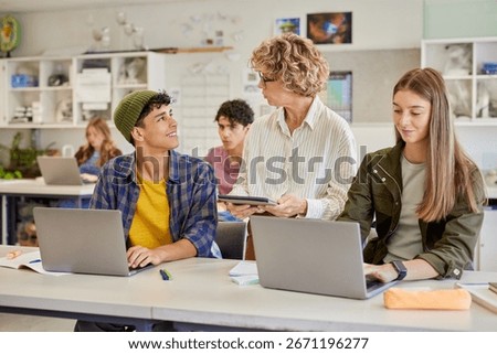 Student talking and smiling at teacher while working on laptop. Instructor holding a digital tablet and interacting with teenagers during digital lesson at secondary school. Professor helping guy.