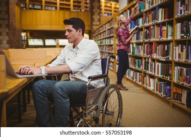 Student In Wheelchair Typing On His Laptop While Woman Searching Books In Library