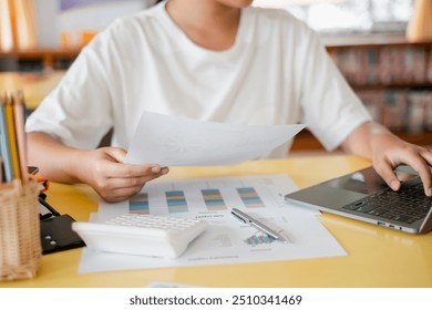 A student in a white shirt analyzing financial data with a laptop and calculator on a yellow desk in a classroom. - Powered by Shutterstock