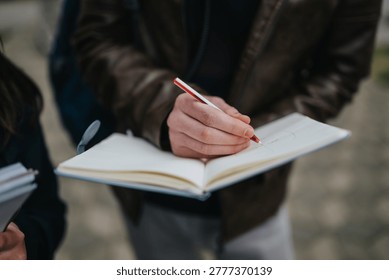A student writes in an open notebook with a red pen during an outdoor study session. Another student holds materials nearby as they learn together on a sidewalk.
