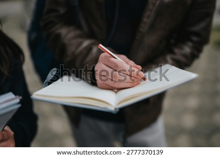 A student writes in an open notebook with a red pen during an outdoor study session. Another student holds materials nearby as they learn together on a sidewalk.
