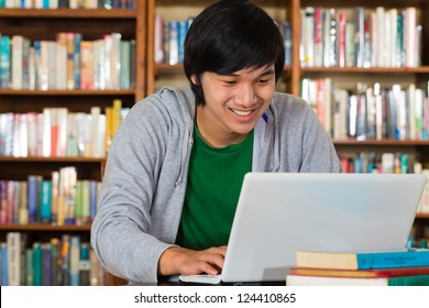 Student - Young Asian man in library with laptop and a pile of books learns
