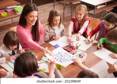 Students boys and girls sitting together around the table in classroom and drawing. With them is their young and beautiful teacher. She teaches children and is smiling - Powered by Shutterstock