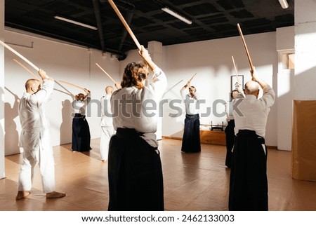 Students clad in white uniforms stand in rows inside a dojo, diligently practicing wooden sword techniques under the guidance of their instructor. Text: 