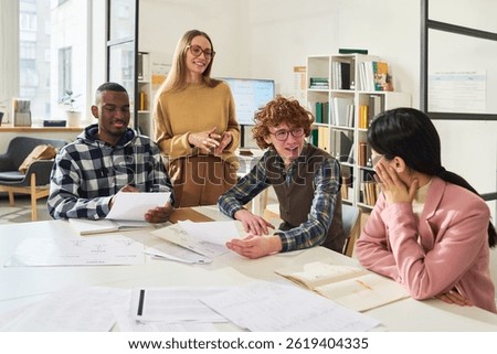 Students engaging in group study session reviewing foreign languages with materials and textbooks around large table. Instructor standing nearby, assisting and smiling