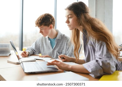students sit at shared desk making notes studying together at university.