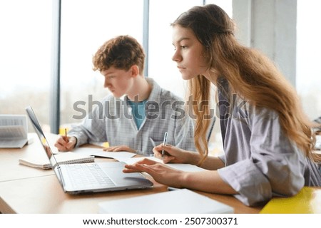 students sit at shared desk making notes studying together at university.