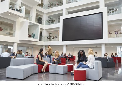 Students sit talking under AV screen in atrium at university