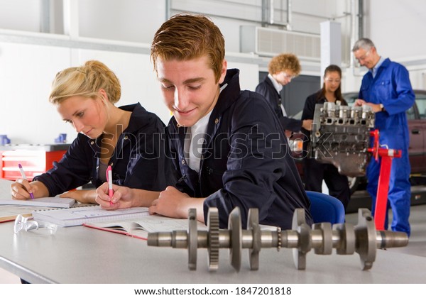 Students sitting on\
the desk studying an auto part kept in the foreground in a\
vocational school of an automotive trade with a trainer and some\
students standing in the\
background