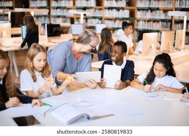 Students and teacher in a library, using tablets and writing. Diverse group of children learning, with a teacher assisting. Engaged in study and technology. Elementary school children at library.