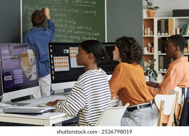 Students working on computers during a lesson with a teacher writing on chalkboard and bookshelf in background. Classroom environment fostering collaborative learning