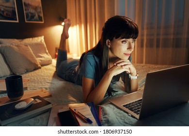 Studing at home online. Pretty young woman using laptop while lying on the bed at night time. - Powered by Shutterstock