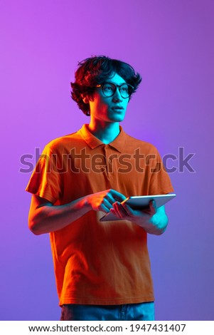 Studing, working. Young man in glasses using tablet isolated on multicolored background in neon light. Concept of human emotions, facial expression, youth culture, digital life. Copy space for ad.