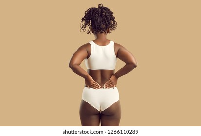 Studio backview shot of beautiful plus size young black woman in underwear standing on solid beige color background and holding hands on her lower back. Concept of body fat, female beauty, and health