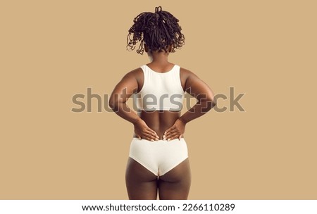 Studio backview shot of beautiful plus size young black woman in underwear standing on solid beige color background and holding hands on her lower back. Concept of body fat, female beauty, and health