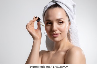 Studio Beauty Photo Portrait Of Lovely Attractive Woman With Head Wrapped In Towel Applying Cosmetic Oil For Facial Skincare. Holding Pipette With Serum Near Clean Face. Cosmetology And Spa