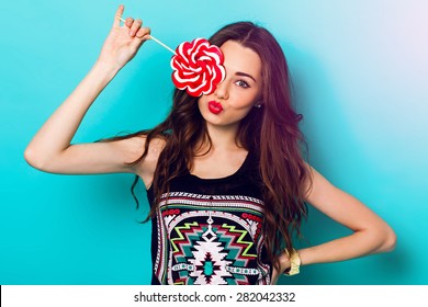 Studio closeup colorful portrait of young sexy funny fashion girl posing on blue wall background in summer style outfit with red lollipop wearing blue jeans and boho t-short.