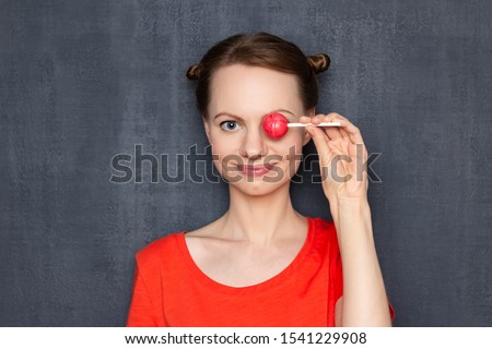 Studio close-up portrait of cheerful happy girl wearing orange T-shirt, with funny hairstyle, covering one eye with pink lollipop, smiling broadly, having fun, fooling around, over gray background