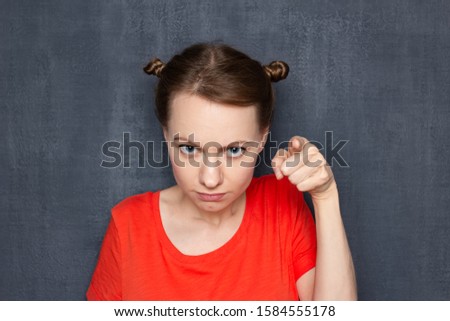 Studio close-up portrait of cute angry dissatisfied girl wearing T-shirt, with funny hairstyle, with frowning face, pointing with index finger at you, scolding and making rebuke, over gray background