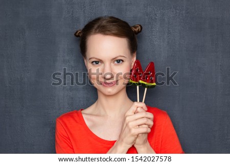 Studio close-up portrait of cute happy pretty woman wearing orange T-shirt, with funny hairstyle, holding two lollipops in hands near head, smiling cheerfully, being in good mood, over gray background