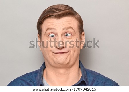 Studio close-up portrait of funny confused blond mature man making crazy goofy face, with crossed eyes, expressing puzzlement, feeling bewilderment. Headshot over gray background