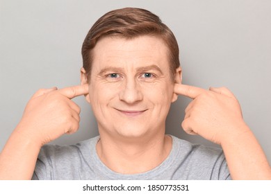 Studio Close-up Portrait Of Happy Blond Mature Man Plugging Ears With Fingers, Not Wanting To Listen To Anyone, Looking Optimistic And Joyful, Smiling Cheerfully. Headshot Over Gray Background