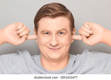 Studio Close-up Portrait Of Happy Blond Mature Man Plugging Ears With Fingers, Not Wanting To Listen To Anyone, Looking Optimistic And Joyful, Smiling Cheerfully. Headshot Over Gray Background