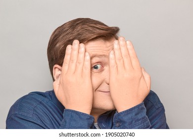 Studio close-up portrait of happy blond mature man covering his face with hands, smiling joyfully, looking with one eye at something interesting, peeking with curiosity. Headshot over gray background
