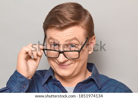 Studio close-up portrait of happy blond mature man looking over his glasses at you, holding glasses frame with hand, smiling cheerfully, with joyful expression on face. Headshot over gray background