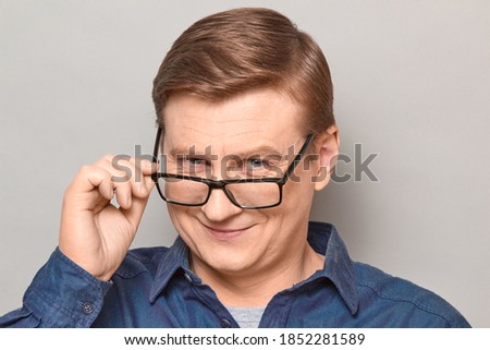 Studio close-up portrait of happy blond mature man looking over his glasses at you, holding glasses frame with hand, smiling cheerfully, with joyful expression on face. Headshot over gray background