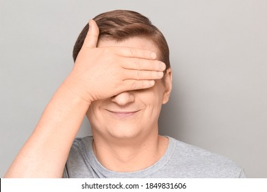 Studio Close-up Portrait Of Happy Optimistic Blond Mature Man Covering Eyes With One Hand, Not Wanting To See Something Or Waiting Surprise, Smiling Cheerfully. Headshot Over Gray Background