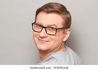 Studio Close-up Portrait Of Joyful Blond Mature Man With Glasses, With Happy Expression On Face, Smiling Cheerfully, Turning Around And Looking Straight Ahead. Headshot Over Gray Background