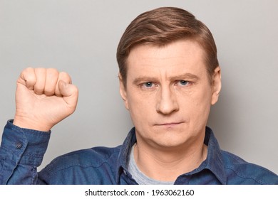 Studio Close-up Portrait Of Serious Blond Mature Man Raising Fist Up, Looking Rebellious And Confident, Protesting, Being Ready To Protect His Rights. Headshot Over Gray Background