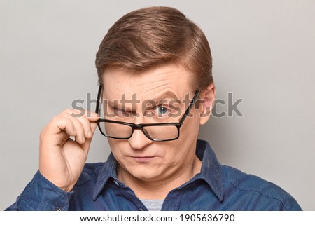 Studio close-up portrait of serious blond mature man looking over his glasses at something interesting, scrutinising something with skeptical strict expression on face. Headshot over gray background