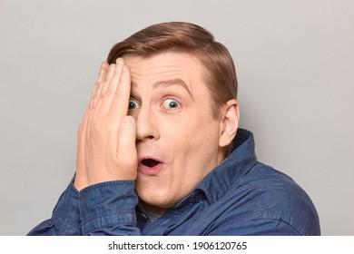 Studio close-up portrait of surprised impressed mature man covering half of his face with hands, looking at something amazing, spying, peeking with curiosity. Headshot over gray background