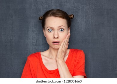 Studio close-up portrait of unhappy sad frightened girl wearing orange T-shirt, with funny hairstyle, touching cheek with hand while being surprised of news or having toothache, over gray background