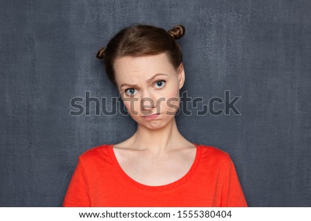 Studio close-up portrait of upset disappointed young woman wearing orange T-shirt, with funny hairstyle, with apologetic expression, being sad and embarrassed about what happened, over gray background