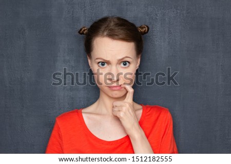 Studio close-up portrait of upset perplexed doubting girl wearing orange T-shirt, with funny hairstyle, holding finger in mouth, regretting about happened, worrying about results, over gray background