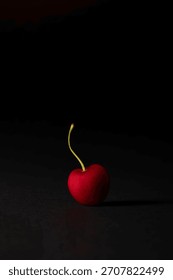 Studio close-up of a single perfect, dark red cherry with a curved stem, isolated against a stark black background. High contrast, minimalist food photography focusing on health and design.
