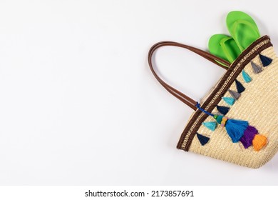 Studio closeup top view shot of green rubber casual unisex slipper in woven weaving wicker rattan handbag decorated with colorful tassel for summer beach sea weekend vacation on white background.