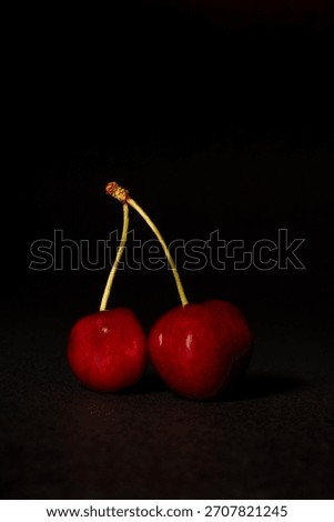 Studio close-up of two perfect, dark red cherries connected by their stems, isolated against a stark black background. High contrast, minimalist food photography focusing on health and design.