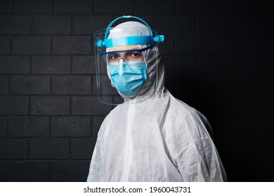 Studio Dark Portrait Of Young Doctor Man Wearing PPE Suit Against Coronavirus And Covid-19. Background Of Black Brick Wall.