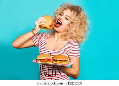 Studio fashion portrait of a beautiful funny young girl on the turquoise background, blonde holding a tray burger is going to bite a hamburger, curly hair, perfect makeup, crazy emotions, fast food